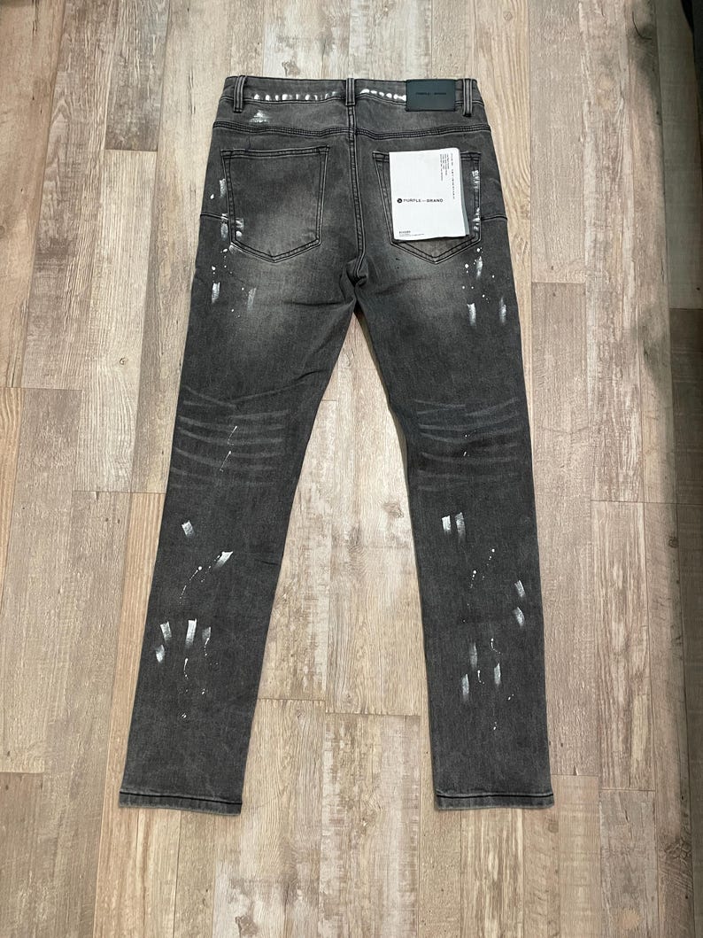 Op de afbeelding: Donkergrijze denim jeans met witte verfspatten. De jeans hebben een klassiek ontwerp met vijf zakken en een rechte pijp. Een klein label is bevestigd aan de achterzak. De jeans liggen plat op een houten oppervlak.