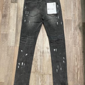 Op de afbeelding: Donkergrijze denim jeans met witte verfspatten. De jeans hebben een klassiek ontwerp met vijf zakken, een knoop- en ritssluiting en een klein label op de achterzak. De jeans liggen plat op een houten oppervlak.
