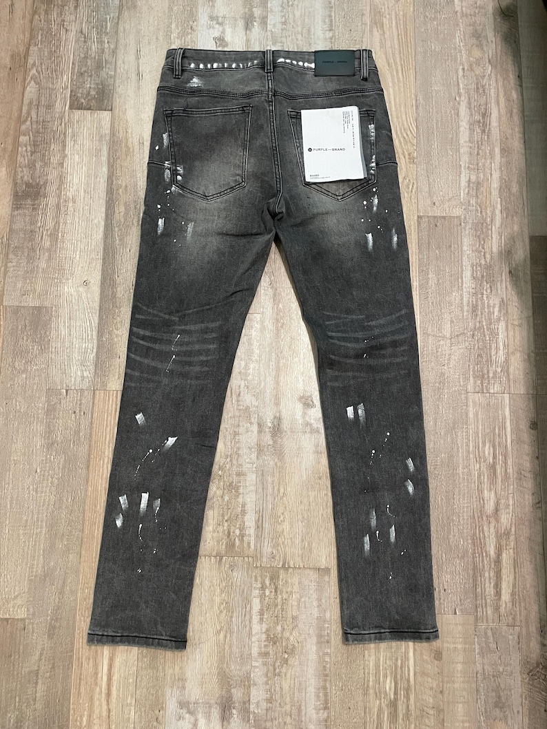 Op de afbeelding: Donkergrijze denim jeans met witte verfspatten. De jeans hebben een klassiek ontwerp met vijf zakken, een knoop- en ritssluiting en een klein label op de achterzak. De jeans liggen plat op een houten oppervlak.