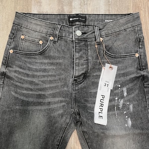 Op de afbeelding: Close-up van een grijze denim jeans met een vervaagde wassing en een distressed look. De jeans heeft een knoopsluiting, vijf zakken en een maat 31 label. De merknaam "PURPLE" is zichtbaar op het label en de jeans.