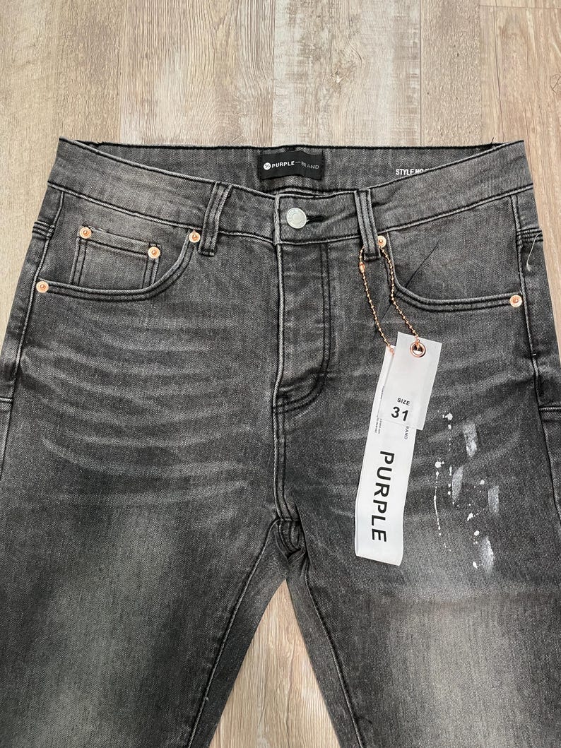 Op de afbeelding: Close-up van een grijze denim jeans met een vervaagde wassing en een distressed look. De jeans heeft een knoopsluiting, vijf zakken en een maat 31 label. De merknaam "PURPLE" is zichtbaar op het label en de jeans.