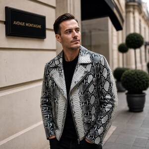 以下が含まれることがあります： 黒と白のヘビ柄の革製ジャケット。ジッパー開閉とラペルカラーが特徴です。ジャケットは黒いシャツの上に着用されています。背景には「AVENUE MONTAIGNE」と書かれた看板があります。