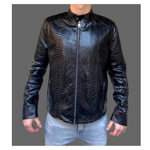 May include: Black leather jacket with a snakeskin texture, featuring a stand-up collar and a full-length zipper. The jacket has long sleeves and a fitted silhouette. The model is wearing light blue jeans. The jacket is a stylish outerwear option.