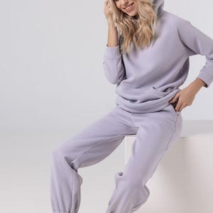 Puede incluir: Un conjunto de sudadera con capucha y pantalón de chándal lila claro. La sudadera tiene un bolsillo delantero y una capucha con cordón. El pantalón de chándal tiene cintura y puños elásticos. La persona lleva zapatillas negras.
