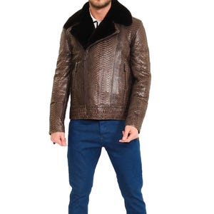 Könnte beinhalten: Eine braune Jacke mit strukturiertem Schlangenledermuster, ausgestattet mit einem breiten schwarzen Kunstfellkragen, langen Ärmeln und einem Frontreißverschluss. Die Jacke wird mit einer blauen Jeanshose kombiniert, was ein komplettes Outfit mit einem deutlichen Kontrast zwischen der strukturierten braunen Außenschicht und dem glatten blauen Stoff der Hose ergibt.