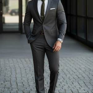 Puede incluir: Un traje gris carbón con camisa blanca, corbata verde oliva y cinturón marrón. El traje presenta una chaqueta de dos botones y pantalones a medida. Se ve un reloj plateado en la muñeca. El conjunto se completa con zapatos negros.