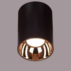 May include: A cylindrical black ceiling light fixture with a gold-colored interior and a bright white light. The light has a modern design, suitable for various interior spaces. The fixture is approximately 6 inches tall.