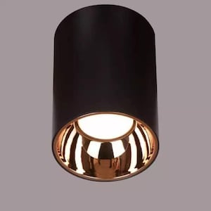 May include: A cylindrical black ceiling light fixture with a gold-colored interior and a bright white light. The light has a modern design, suitable for various interior styles. The fixture is approximately 6 inches tall.