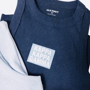 Könnte beinhalten: Ein dunkelblaues geripptes Tanktop mit einem hellblauen quadratischen Aufnäher, der den weißen Text KAPPA KAPPA GAMMA trägt. Das innere Nackenetikett zeigt OLD NAVY Größe XS. Ein hellblaues geripptes Kleidungsstück ist unter dem Tanktop sichtbar.