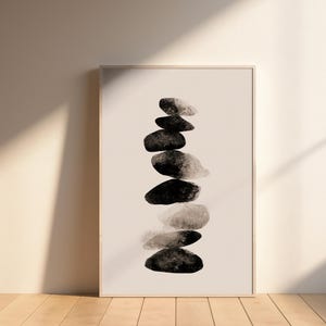 May include: A framed art print featuring a stack of seven abstract stones in shades of black and gray. The artwork is displayed against a neutral background, leaning against a wall. The print is in a light wooden frame.