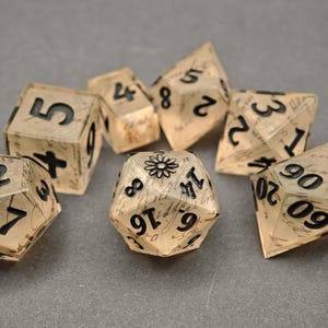 May include: A set of seven polyhedral dice made of translucent beige resin with black ink numbers and a cursive script pattern embedded inside. The twenty-sided die features a small black flower icon in place of the number twenty.