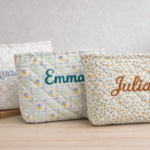 Könnte beinhalten: Drei gesteppte Kosmetiktaschen mit den gestickten Namen: Ava, Emma und Julia. Die Taschen weisen florale und geometrische Muster in sanften Pastellfarben auf. Die Taschen sind aus gestepptem Stoff gefertigt und für Make-up oder andere kleine Gegenstände konzipiert.
