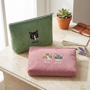 Puede incluir: Un estuche de pana verde salvia con cremallera que presenta una cara de gato bordada en blanco y negro con el nombre Buddy, y un estuche de pana rosa empolvado con las caras bordadas de los gatos Milo y Luna con sus nombres debajo.