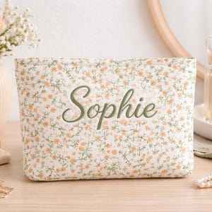 Könnte beinhalten: Kosmetiktasche mit floralem Muster und dem Namen "Sophie" in Grün gestickt. Die Tasche hat einen cremefarbenen Hintergrund mit kleinen orangefarbenen und grünen Blumen. Sie steht auf einer Holzoberfläche, mit Parfümflaschen und Blumen im Hintergrund.