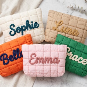 Op de afbeelding: Vijf gewatteerde make-up tassen in crème, beige, oranje, roze en groen. Elke tas heeft een naam geborduurd in een contrasterende kleur: Sophie, Luna, Belle, Emma en Grace. De tassen liggen op een lichtgekleurd oppervlak.