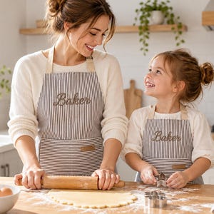 May include: Two matching grey and white striped aprons featuring the word Baker embroidered in cursive script on the chest. Each apron includes a small rectangular patch on the front pocket and beige neck straps, worn over white long-sleeve shirts.