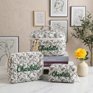 Könnte beinhalten: Ein Set aus drei gesteppten Kosmetiktaschen mit einem schwarz-weißen Blumenmuster. Jede Tasche trägt den Namen "Charlotte" in Grün gestickt. Die größte Tasche hat einen Griff und Quasten-Reißverschlussanhänger.