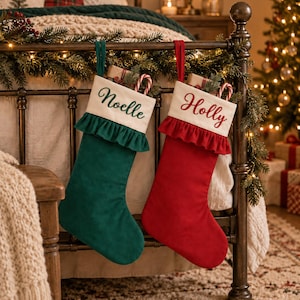 Pode incluir: Duas meias de Natal penduradas numa estrutura de cama de metal. Uma meia é verde-escura com um punho branco bordado com o nome Noelle em verde. A outra é vermelha com um punho branco bordado com o nome Holly em vermelho. Ambas têm folhos.