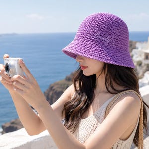 Può includere: Un cappello a secchiello all'uncinetto di colore viola brillante con la parola Lovely ricamata in corsivo bianco sul davanti. Il cappello ha un aspetto intrecciato e testurizzato con tesa spiovente e corona arrotondata in filato spesso.