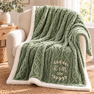 May include: A sage green throw blanket with a textured cable knit pattern and a white sherpa fleece lining. The blanket features an embroidered design with the text R + M and the year 2026, framed by two curved leafy branches.