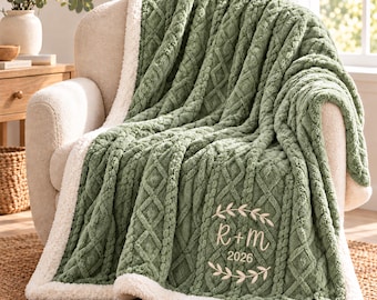 Op de afbeelding: Een saliegroene plaid met een getextureerd kabelgebreid patroon en een witte sherpa fleece voering. De deken is voorzien van een geborduurd ontwerp met de tekst R + M en het jaartal 2026, omlijst door twee gebogen bladeren.