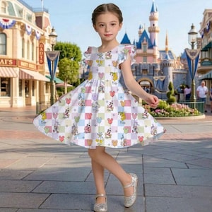 Puede incluir: Un vestido blanco con un colorido estampado de Mickey Mouse y globos. El vestido tiene mangas cortas con bordes con volantes y una falda acampanada. La ni&ntilde;a lleva zapatos plateados con purpurina. El fondo muestra un parque tem&aacute;tico.