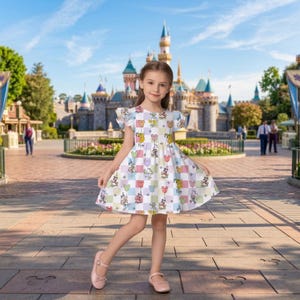 Puede incluir: Un vestido blanco con un estampado de cuadros de colores con personajes de dibujos animados. El vestido tiene mangas cortas y una falda acampanada. El fondo muestra un castillo y un d&iacute;a soleado.