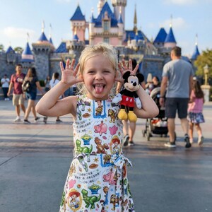 May include: A young child wearing a white dress with a colorful Toy Story print, holding a Mickey Mouse plush toy. The child is smiling and sticking out their tongue in front of a castle. The dress has a bow at the waist.
