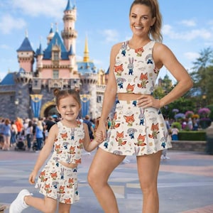 Op de afbeelding: Bijpassende cr&egrave;mekleurige crop tops en rokken met een cartoon vos en konijn print. De outfits hebben de woorden "Hustle" en "Sly!". Een vrouw en een kind dragen de outfits voor een kasteel.