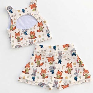 Op de afbeelding: Een cr&egrave;mekleurige tweedelige outfit met een cartoon vos en konijn print. De top is een crop top met een sleutelgat rug, en de shorts hebben een bijpassend ontwerp. De woorden "HUSTLE" en "SLY!" zijn op de stof gedrukt.