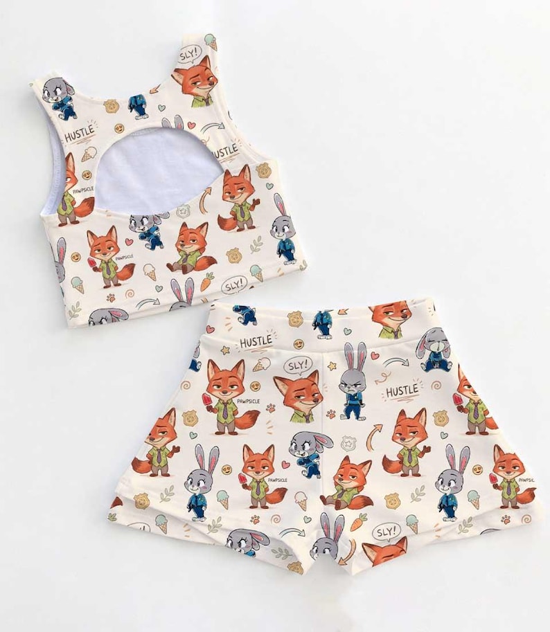 Op de afbeelding: Een cr&egrave;mekleurige tweedelige outfit met een cartoon vos en konijn print. De top is een crop top met een sleutelgat rug, en de shorts hebben een bijpassend ontwerp. De woorden "HUSTLE" en "SLY!" zijn op de stof gedrukt.