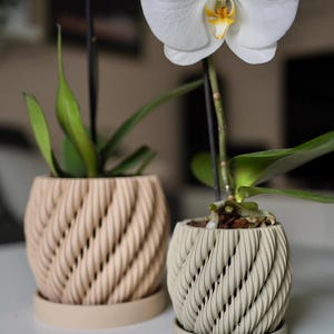 May include: Two cylindrical plant pots with a textured, spiraling rope-like pattern on the exterior. One pot is a light beige color, and the other is a slightly darker tan shade. Each pot sits on a matching circular saucer, with one containing a white orchid.