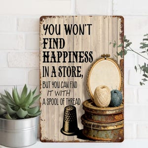 Puede incluir: Un letrero metálico rústico con el texto "YOU WON'T FIND HAPPINESS IN A STORE, BUT YOU CAN FIND IT WITH A SPOOL OF THREAD." El letrero presenta un dedal, un bastidor de bordado y carretes de hilo. El fondo es un diseño de madera desgastada.