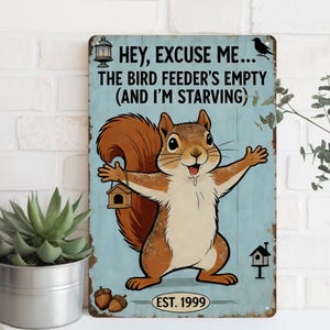 Op de afbeelding: Een metalen bord met een cartoon eekhoorn die een vogelvoederbak vasthoudt. Het bord heeft de tekst "HEY, EXCUSE ME... THE BIRD FEEDER'S EMPTY (AND I'M STARVING)" en "EST. 1999". De eekhoorn is bruin en wit.