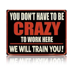 Op de afbeelding: Een rechthoekig metalen bord met een zwarte achtergrond en een rode rand. De tekst in wit en rood luidt: YOU DON'T HAVE TO BE CRAZY TO WORK HERE WE WILL TRAIN YOU! Het bord heeft vier kleine gaatjes in de hoeken voor bevestigingsdoeleinden.