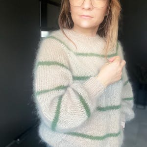 May include: A beige, oversized sweater with horizontal green stripes. The sweater has long sleeves and a crew neck. The material appears soft and fluffy. The person in the image is wearing glasses.