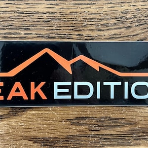 May include: A rectangular black sticker with the words "PEAK EDITION" in orange and white. Above the text is an orange mountain range graphic. The sticker is on a wooden surface.