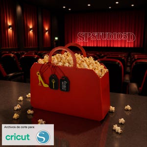 Puede incluir: Un recipiente rojo de palomitas de maíz lleno de palomitas, con dos asas y etiquetas decorativas. El recipiente está sobre una superficie oscura, con granos de palomitas esparcidos. El fondo muestra una sala de cine con cortinas rojas y asientos.