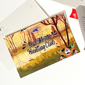 May include: A grey and white Nintendo Zapper light gun with a black cord. The Zapper is on top of a postcard with a cartoon illustration of a woodland scene and the text "Fowl Mouth Hunting Club".
