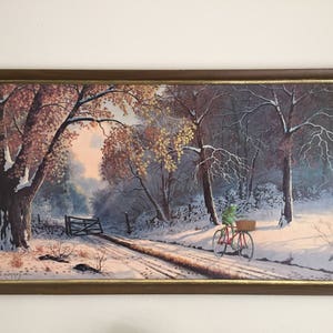May include: Framed landscape painting depicting a snowy road through a forest. A red bicycle with a basket is ridden by a green frog. The painting has a brown frame with a gold inner edge. The artwork is signed in the lower left corner.