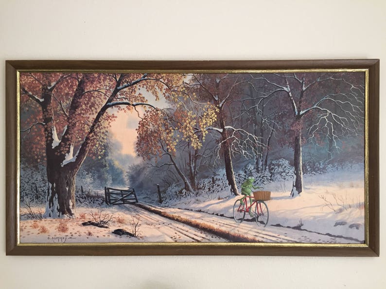May include: Framed landscape painting depicting a snowy road through a forest. A red bicycle with a basket is ridden by a green frog. The painting has a brown frame with a gold inner edge. The artwork is signed in the lower left corner.