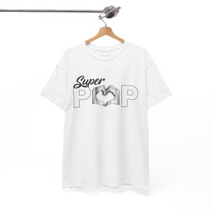May include: White t-shirt with the words "Super Pop" in a black script font. The "O" in "Pop" is a heart shape formed by two hands. The shirt is hanging on a wooden hanger.