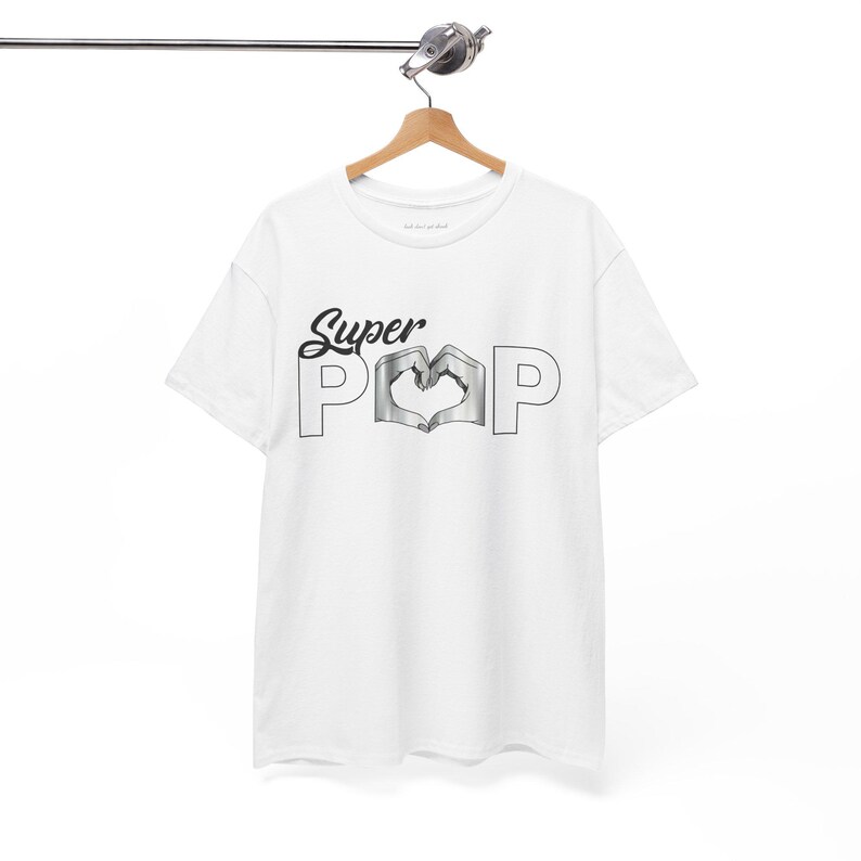 May include: White t-shirt with the words "Super Pop" in a black script font. The "O" in "Pop" is a heart shape formed by two hands. The shirt is hanging on a wooden hanger.