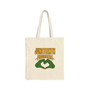 May include: Cream-colored tote bag with the words "JUNETEENTH FREEDOM" in orange and yellow. Below the text is a green heart shape made of hands, with a chain design. The bag has long handles.