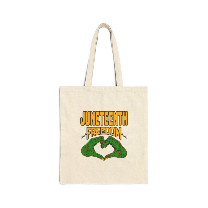 May include: Cream-colored tote bag with the words "JUNETEENTH FREEDOM" in orange and yellow. Below the text is a green heart shape made of hands, with a chain design. The bag has long handles.