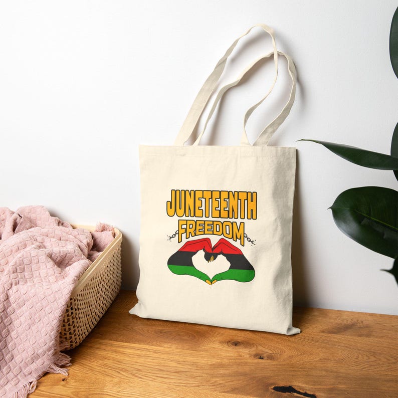 May include: Cream-colored tote bag with the words "Juneteenth Freedom" in gold and black. The design includes a heart shape formed by hands in the colors of the Pan-African flag. The bag has long handles.