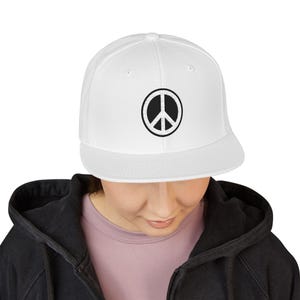 May include: A white snapback cap featuring a circular black and white embroidered peace sign emblem centered on the front panel. The hat has a structured crown, a flat brim, and small ventilation eyelets positioned around the top of the crown.