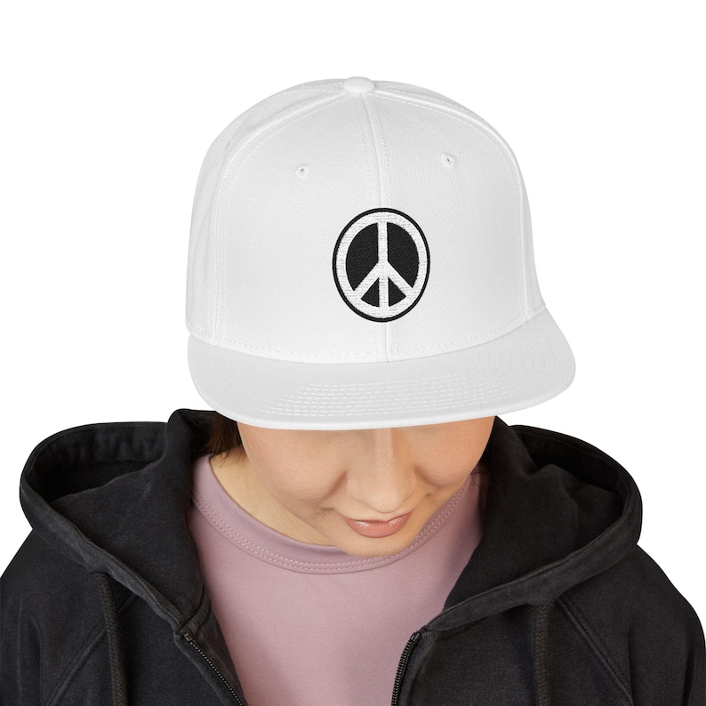 May include: A white snapback cap featuring a circular black and white embroidered peace sign emblem centered on the front panel. The hat has a structured crown, a flat brim, and small ventilation eyelets positioned around the top of the crown.