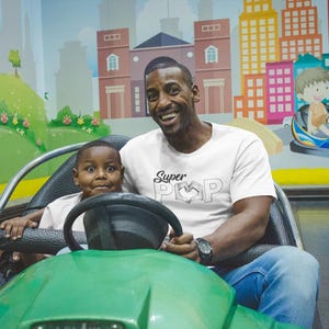 May include: A father and child are in a green bumper car at an amusement park. The father wears a white t-shirt with "Super Pop" printed on it. The background features a colorful mural of a city and a child in a blue bumper car.