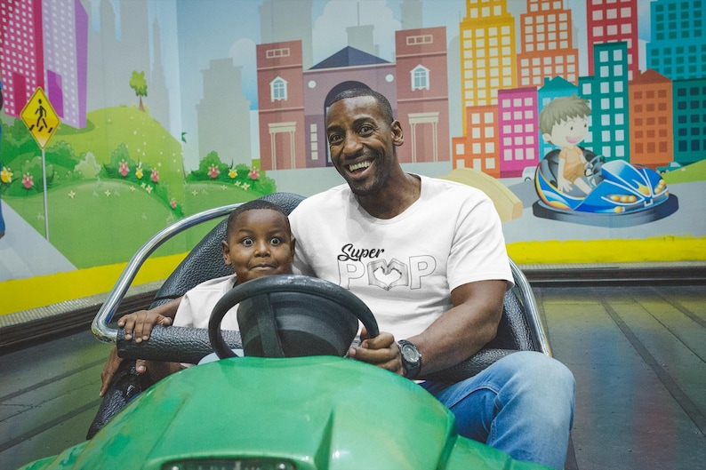 May include: A father and child are in a green bumper car at an amusement park. The father wears a white t-shirt with "Super Pop" printed on it. The background features a colorful mural of a city and a child in a blue bumper car.